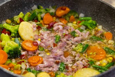 Renkli bir sebze ve pastırma karışımı bir tavada hazırlanıyor. Çanağın malzemelerinin statik yakın çekimi, yapay ışık altında çekiliyor..