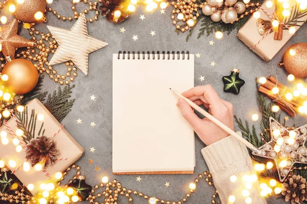 Notepad and writing female hand with holiday decorations in green and golden colors on grey concrete backdrop, copyspace. Christmas and New Year planning flatlay concept.