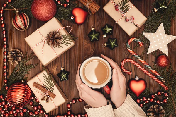 圣诞和新年乡村传统作曲 热咖啡杯在女性的手 在深褐色的旧木板上装饰精美的雪松枝条 礼品盒 星和藤条棒棒糖 — 图库照片#