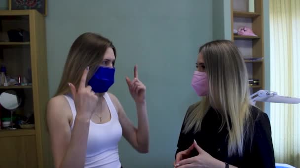 La mujer joven discute el procedimiento con el cosmetólogo en el abrigo negro. Ambos en máscaras — Vídeos de Stock