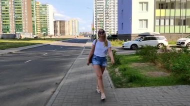 Güneş gözlüklü sarışın kadın kameraya doğru yürür, durur ve güneş gözlüğü takar. Yaz