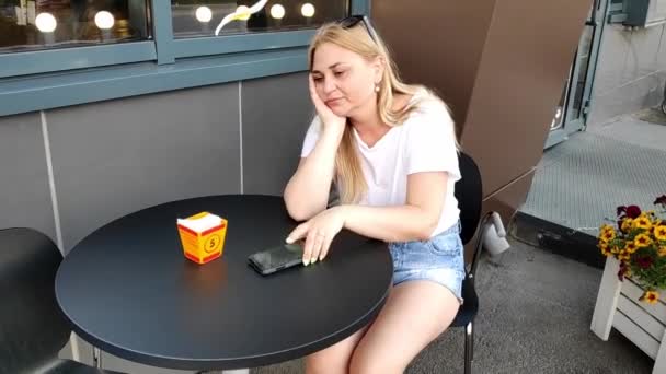 Jonge blonde vrouw wacht op een telefoontje aan de cafe tafel, neemt gelukkig haar telefoon op — Stockvideo