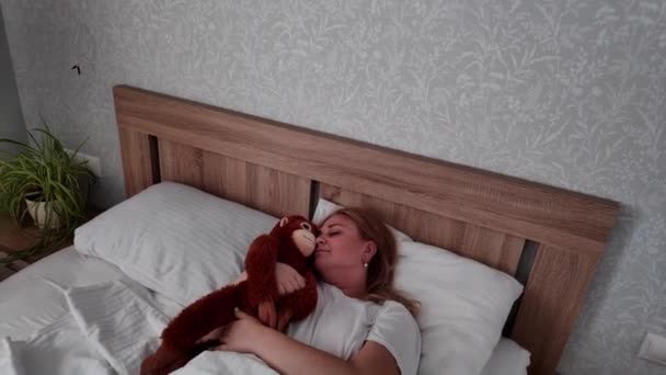 Handheld mulher loira branca acorda na cama com brinquedo de pelúcia macaco, estica — Vídeo de Stock