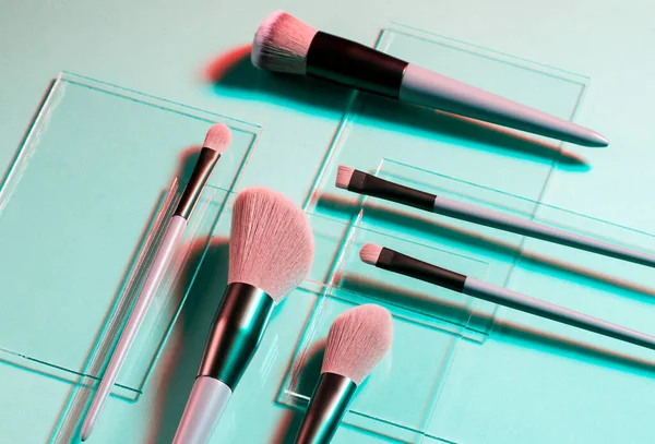 Flat lay of makeup brushes on pieces of glass on the light blue background close up. Neon light. Cosmetic products