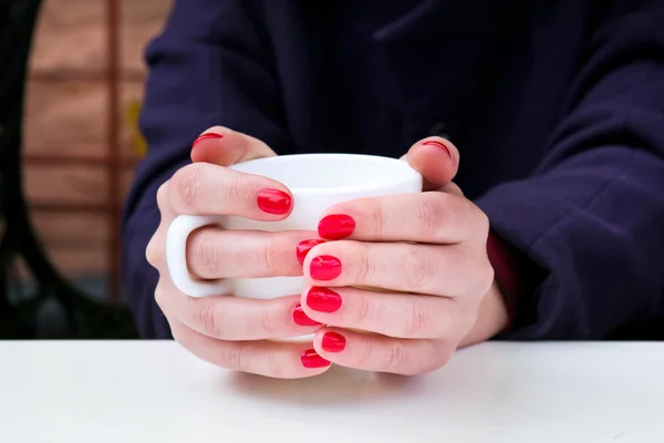 Mujer con esmalte de uñas rojo sosteniendo taza blanca en un café al aire libre — Foto de Stock