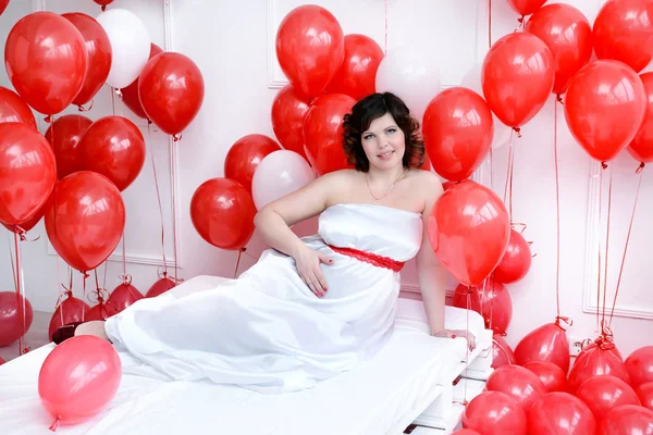 Bir yatakta döşeme ve bir karın çevresinde kırmızı balonlar tutan genç hamile kadın
