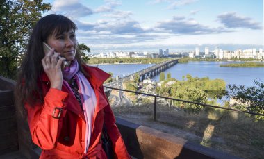 Yan görünüm fotoğrafı. Telefonla konuşan, açık havada güneşli bir sonbahar gününde şehir manzarasına bakan tatlı bir kız.