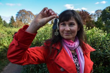 Kırmızı paltolu güzel esmer bir kız elinde yeni evin anahtarlarını tutuyor, doğal arka plana karşı. 