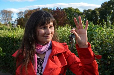 Kırmızı paltolu güzel esmer bir kız elinde yeni evin anahtarlarını tutuyor, doğal arka plana karşı. 