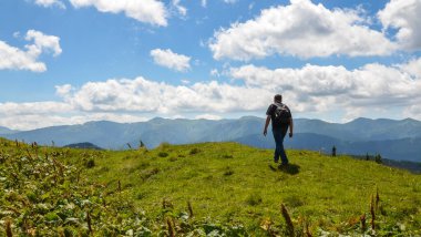 Sırt çantalı genç yürüyüşçünün arka görüntüsü çayırda yürürken bir dağ yolu güneşli bir dağın keyfini çıkarıyor. 