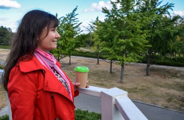 Açık havada, kırmızı ceketli, tek kullanımlık kahve fincanı tutan ve şehir doğasına karşı gülümseyen güzel esmer kadının portresi. Kahve hazır. 