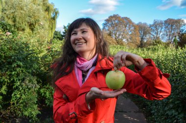 Kırmızı ceketli güzel, gülümseyen esmer kız avucunun içinde taze yeşil elma tutuyor. Yiyecek, meyve ve sağlıklı beslenme