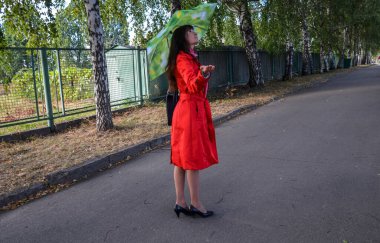 Uzun saçlı esmer bir kızın güzel portresi. Kırmızı ceketli, yeşil şemsiyeli, yağmurun yağıp yağmadığını bulmaya çalışan.