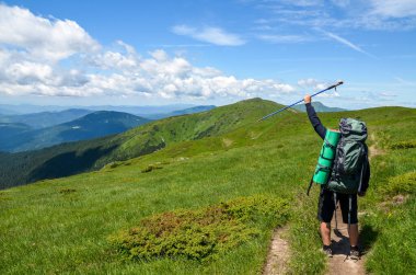 Sırt çantalı turist, yeşil orman ve güzel tepelerin doğal manzarasına karşı elini kaldırarak kameraya geri dönüyor..