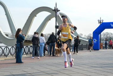 Sarı kolsuz gömlek ve beyaz çorap giyen erkek koşucu, seyircilerin fotoğraf ve tezahürat çekerken nehir kıyısındaki sahil yolunda yarışıyor.