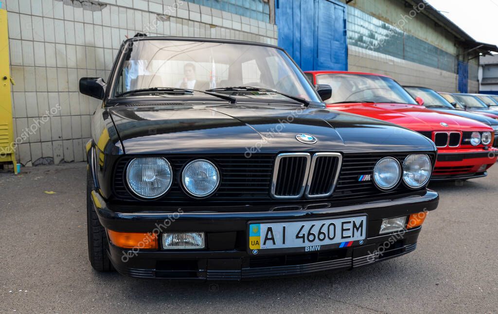 Black BMW E30 takes the foreground at an outdoor car show, with a line of classic cars including a red BMW in the background; a stylish, vintage automotive display