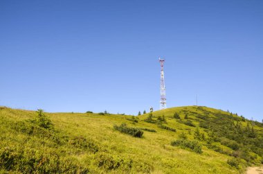 İletişim kulesi açık mavi bir gökyüzünün altında çimenli bir yamaçta duruyor. Tepe yeşil çimlerle ve birkaç alçak çalılarla kaplı, yamaçta birkaç ağaç var.