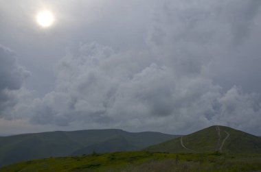 Moody landscape of rolling green hills under dramatic, overcast sky. The sun shines through the thick clouds, creating bright white orb in the upper left corner. A faint path winds up one of the hills