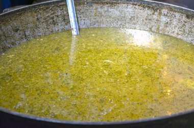 Large metal cauldron holds a bubbling, green-hued soup, with a ladle resting inside. The surface of the liquid shows bits of herbs and ingredients, hinting at a savory, hearty dish