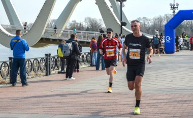 İki koşucu nehir kenarındaki bir gezinti alanında yarışırken, seyirciler arka planda belirgin beyaz bir kemer köprüsü ve canlı bir şehir ortamı ile izliyorlar.