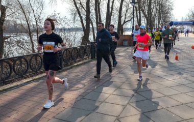 Bir grup koşucu nehir kenarındaki sahil şeridi boyunca gündüz yarışına katılıyor. Atletizmdeki enerjik atletler, seyirciler ve sakin nehir zeminiyle ileri doğru itiliyor.