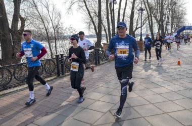 Bir grup koşucu nehir kenarındaki sahil şeridi boyunca gündüz yarışına katılıyor. Atletizmdeki enerjik atletler, seyirciler ve sakin nehir zeminiyle ileri doğru itiliyor.