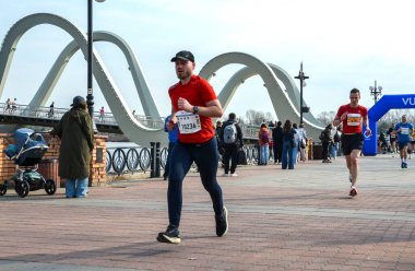 İki koşucu nehir kenarındaki bir gezinti alanında yarışırken, seyirciler arka planda belirgin beyaz bir kemer köprüsü ve canlı bir şehir ortamı ile izliyorlar.