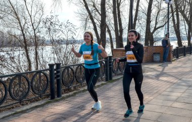 Bir grup koşucu, asfalt bir gezinti alanı boyunca güneşli bir nehir kıyısındaki yarışa katılıyor. Hareketli adımlar ve atletik giysiler spor, takım çalışması ve açık hava eğlencesi.