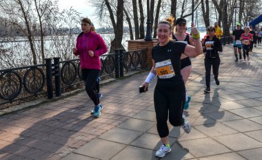 Bir grup koşucu, asfalt bir gezinti alanı boyunca güneşli bir nehir kıyısındaki yarışa katılıyor. Hareketli adımlar ve atletik giysiler spor, takım çalışması ve açık hava eğlencesi.
