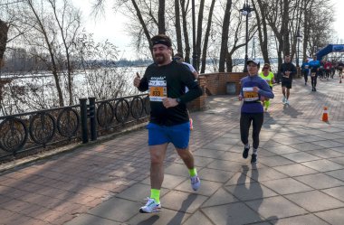 Bir grup koşucu, asfalt bir gezinti alanı boyunca güneşli bir nehir kıyısındaki yarışa katılıyor. Hareketli adımlar ve atletik giysiler spor, takım çalışması ve açık hava eğlencesi.