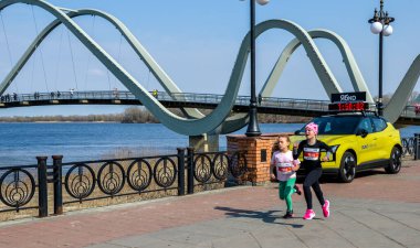 İki genç kız açık hava koşusu sırasında nehir kenarında bir gezinti güvertesinde yarışıyorlar. Sarı destek aracı ve dijital zamanlayıcı rekabetçi sahneye enerji ve hız ekler