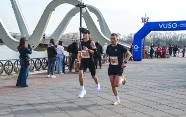 Koşucular dramatik, kavisli köprünün altında nehir kenarındaki gezinti boyunca yarışırken, izleyiciler spor, spor ve açık hava aktivitelerinin canlı görüntülerini izliyorlar.