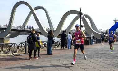 Koşucular dramatik, kavisli köprünün altında nehir kenarındaki gezinti boyunca yarışırken, izleyiciler spor, spor ve açık hava aktivitelerinin canlı görüntülerini izliyorlar.