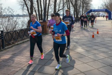Nehir kıyısındaki bir kaldırım boyunca koşan kadınlar renkli atletizm kıyafetleriyle sürüye önderlik ediyorlar. Diğer koşucular ve umutlu destekçiler yakınlarda