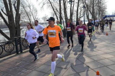 Nehir kıyısındaki bir kaldırım boyunca koşan kadınlar renkli atletizm kıyafetleriyle sürüye önderlik ediyorlar. Diğer koşucular ve umutlu destekçiler yakınlarda