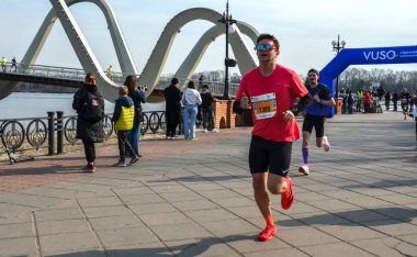 Koşucular dramatik, kavisli köprünün altında nehir kenarındaki gezinti boyunca yarışırken, izleyiciler spor, spor ve açık hava aktivitelerinin canlı görüntülerini izliyorlar.