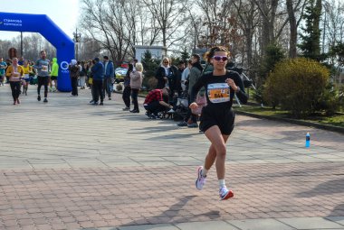 Kadın sporcu güneşli bir parkta koşuyor. Odaklanma ve atletizm gösteriyor.