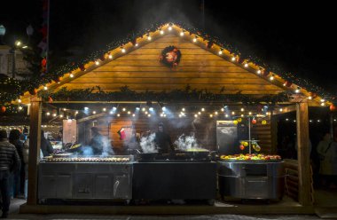 Ahşap gıda tezgahı gece yılbaşı ışıklarıyla aydınlık olur. Satıcılar yiyecek hazırlarken, büyük yemek yüzeylerinden buhar yükseliyor ve festival atmosferi yaratıyor.