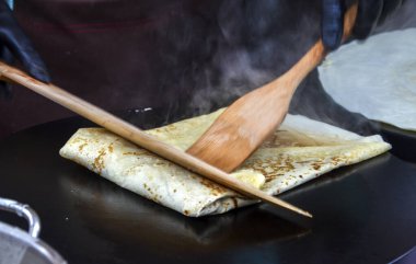 Siyah eldiven giyen şef, ahşap spatula kullanarak büyük, siyah bir ızgaranın üzerine krep katlıyor. Taze pişmiş hamur işlerinden buhar yükselir