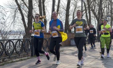 Çoğunluğu kadın olmak üzere bir grup koşucu, güneşli bir park yolunda önlükleriyle koşuyor, spor yapıyor, takım çalışması yapıyor ve nehir kenarındaki bir yol boyunca yapılan ortak bir yarış sırasında neşeyi paylaşıyor.