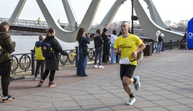 Erkek koşucu nehir kenarında seyircilerle birlikte yarışır ve arka planda çarpıcı bir kemer köprüsü, açık hava fitness ve rekabetçi ruhu yakalar.