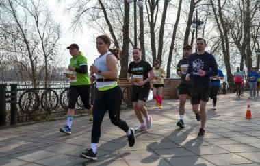 Renkli atletizm kıyafetleri içindeki koşucular rıhtım boyunca yarışır, önlükler görünür.