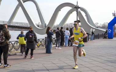 Erkek koşucu nehir kenarında seyircilerle birlikte yarışır ve arka planda çarpıcı bir kemer köprüsü, açık hava fitness ve rekabetçi ruhu yakalar.