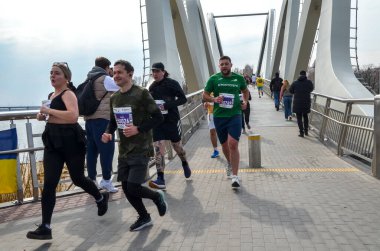 Bir grup koşucu gündüz yarışı sırasında şık şehir köprüsünden geçiyor. Katılımcılar atletizm kıyafeti, takım çalışması, dayanıklılık ve açık hava spor etkinliği giyiyorlar.