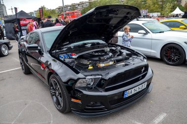 Parlak siyah Ford Mustang kaputu açık, güçlü V8 motorunu ortaya çıkarıyor. Araba spor bir tasarıma, siyah çoklu tekerlere ve bir ön ayırıcıya sahip.