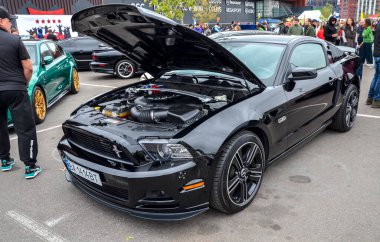 Parlak siyah Ford Mustang kaputu açık, güçlü V8 motorunu ortaya çıkarıyor. Araba spor bir tasarıma, siyah çoklu tekerlere ve bir ön ayırıcıya sahip.