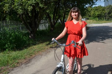 Parlak kırmızı benekli elbiseli kadın güneşli bir park yolunda beyaz bir bisikletin yanında duruyor. Yeşillik ağaçlar ve bir çit çerçevesi Sakin bir açık hava manzarası