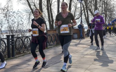 Önlük numaralarına sahip bir grup kadın nehir kenarındaki ağaçlık bir gezinti güvertesinde koşuyor, gündüz açık hava yarışı sırasında enerji, takım çalışması ve sağlıklı yaşam sergiliyorlar.