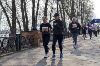 Önlük numaralarına sahip bir grup kadın nehir kenarındaki ağaçlık bir gezinti güvertesinde koşuyor, gündüz açık hava yarışı sırasında enerji, takım çalışması ve sağlıklı yaşam sergiliyorlar.