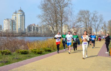 Nehir kenarındaki güneşli şehir parkında çeşitli koşucular yarışırken, arka planda şehir silüeti yer alıyor. Parlak giysiler içindeki enerjik sporcular canlı yaratıyor, yarış atmosferi yaratıyorlar.